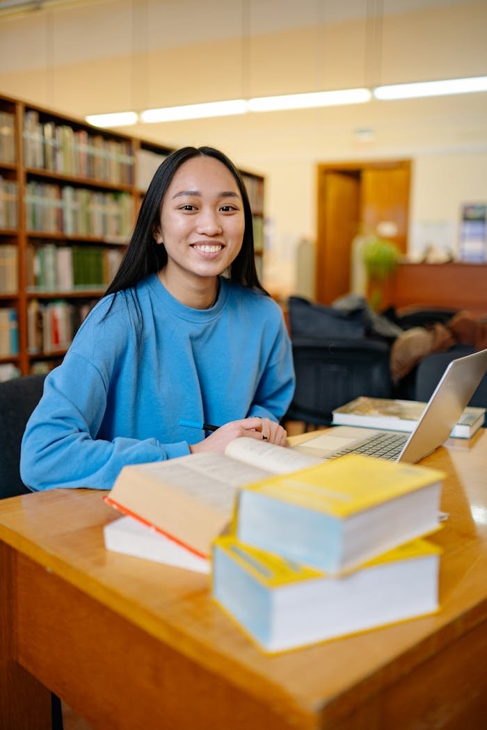 A young woman student sitting in a library with books and a laptop, smiling while studying.